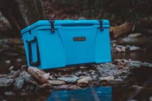 teal kenai cooler sitting on river bank with reflection of cooler on water