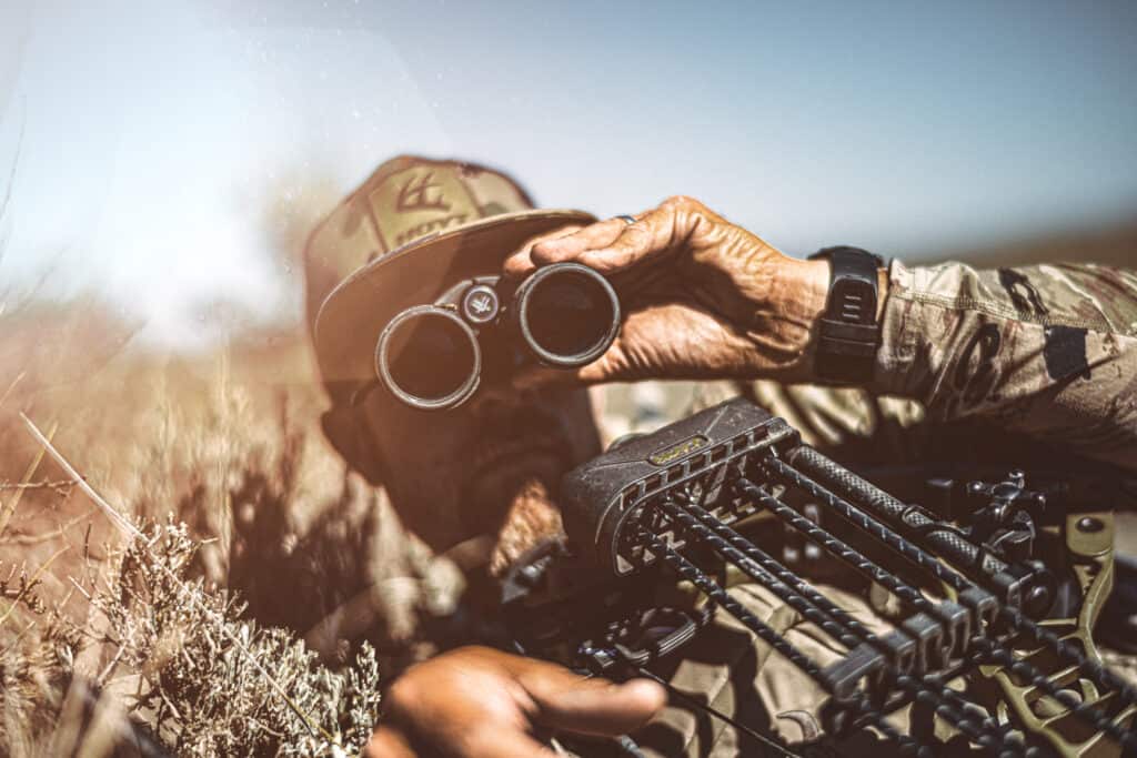 Antelope Hunter Using Binoculars Sitting In Ground Blind With Bow Resting On Chest