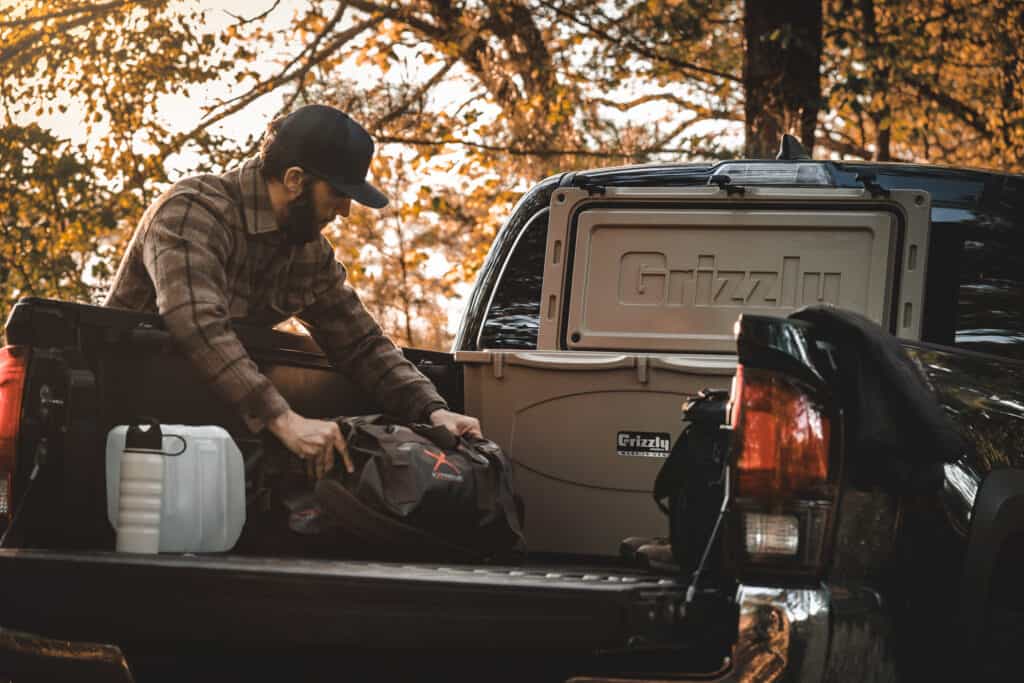 Grizzly 75 Cooler With Lid Open Sitting In Back Of Pick Up With Man Reaching Into Back Of Truck Bed With Hunting Gear.