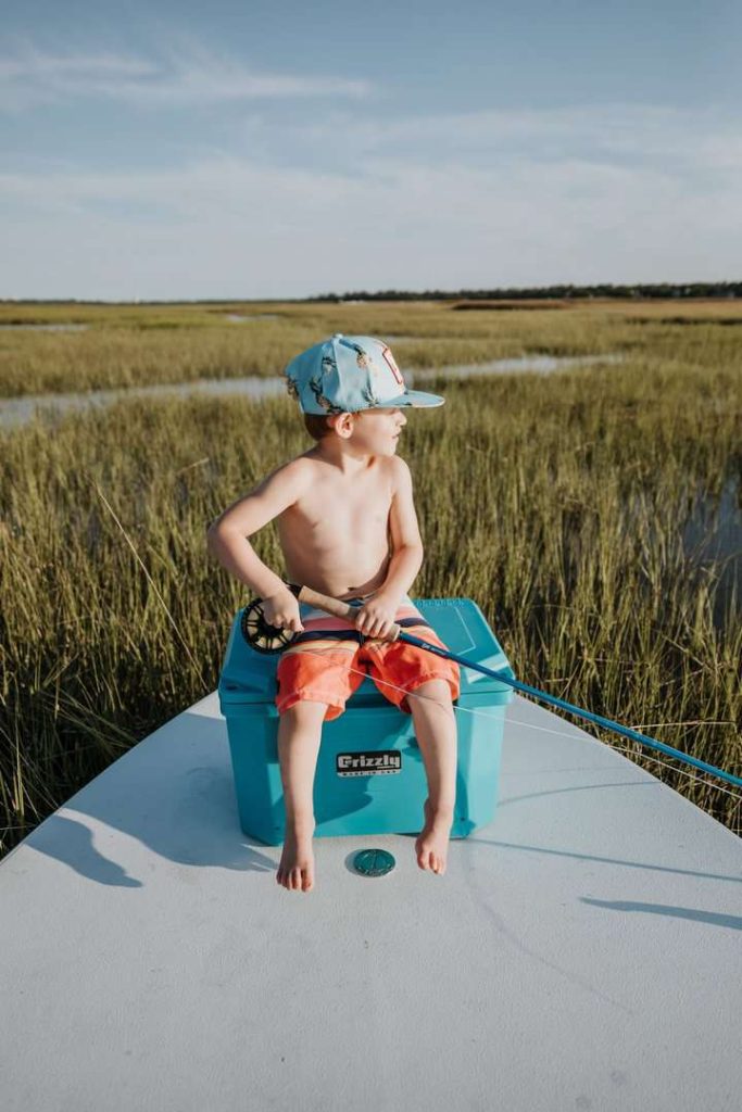 Boy Wearing A Hat Sitting On Teal Grizzly 20 Cooler Holding A Fly Fishing Rod Looking To The Side